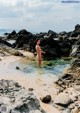 A naked woman standing in the water on a rocky beach.